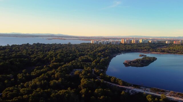 Aerial drone orbit around a lake in Albufera Natural Park, Valencia. Wetlands and sandy dunes with Valencia city skyline in the distance at sunset.