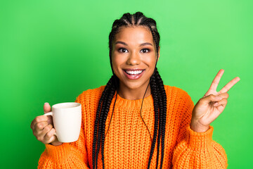 Young woman with braided hair wearing an orange sweater holds a mug and flashes a peace sign against a green background