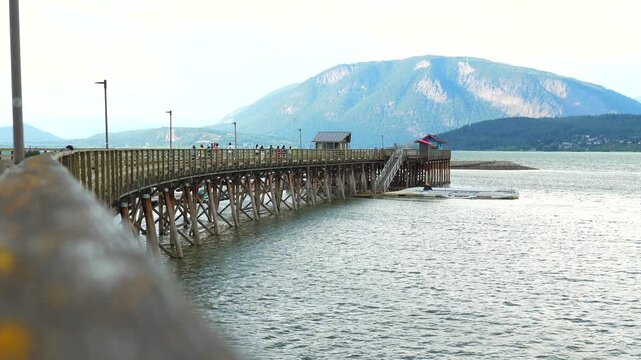 Wooden pier stretching into serene shuswap lake, showcasing scenic mountain vistas and tourists enjoying panoramic views of mount ida in salmon arm, british columbia