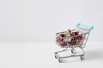 Mini shopping cart with Christmas baubles ornaments, blurred white background. Festive holiday discount concept. Christmas shopping, seasonal sales. Winter retail promotions, gift buying themes banner