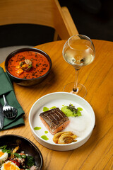 A beautifully plated salmon fillet garnished with green puree sits next to a warm bowl of rich soup and a glass of crisp white wine. The setting is intimate and inviting