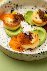 Succulent scallops rest on a bed of bright green puree, adorned with a crunchy topping and a drizzle of vibrant sauce, showcasing culinary art in a cozy evening setting