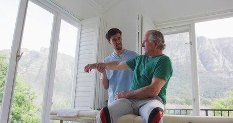 Therapist in blue scrubs supporting senior patient in green tee with knee braces in therapy room