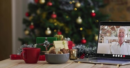 Chatting senior woman on laptop in white shirt, waving at holiday home with cookies, copy space