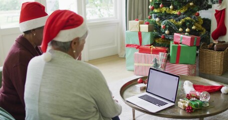 Sitting seniors wearing red Santa hats, sharing open laptop at home with decorated tree and gifts