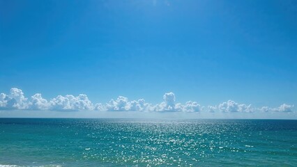 Fototapeta premium A serene ocean view with a bright blue sky, scattered clouds, and sunlight reflecting off the water.