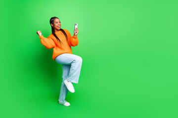 Young woman in orange sweater dancing with phone against bright green background