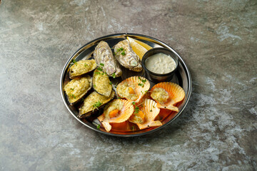 A tempting selection of baked oysters and scallops sits on a metallic plate, drizzled with herbs and accompanied by a zesty dipping sauce, creating a mouthwatering experience