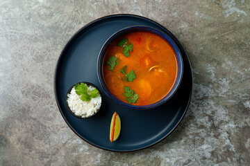 A vibrant bowl filled with steaming soup sits on a dark plate, accompanied by a small bowl of rice and a lime wedge. Fresh cilantro adds a pop of color