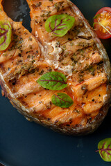 Richly grilled salmon steak is adorned with delicate green herbs and bright red cherry tomatoes, creating a feast for the senses on a dark plate