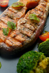A beautifully grilled salmon fillet lies on a black plate, garnished with fresh basil. Vibrant cherry tomatoes and steamed broccoli add a burst of color, showcasing a healthy dish