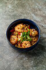 A bowl brimming with flavorful shrimp and colorful vegetables sits on a textured surface. The golden noodles are topped with fresh green onion, creating a deliciously inviting dish