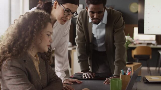 Tilt shot of curly haired woman using laptop to demonstrate report to her biracial colleagues who gathered around her desk and looking at laptop screen