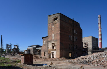 An abandoned factory with pipes, tractor, dilapidated brick buildings and pieces of walls under a clear blue sky