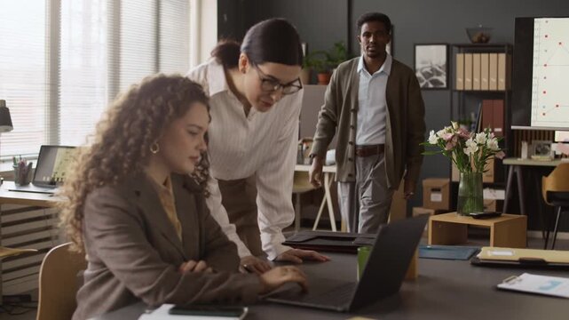 African American man coming to two Caucasian women who looking at laptop screen and checking report. They paying attention to him and smiling, asking for help