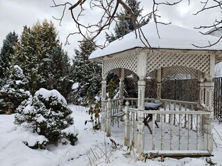 White gazebo with a bench and a bird bath in the snow. The gazebo is surrounded by trees and bushes