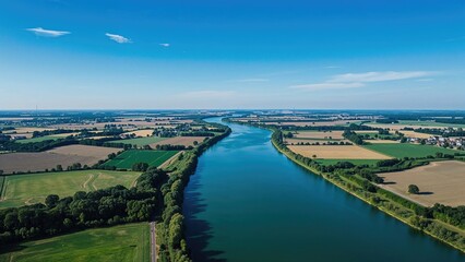 Fototapeta premium River landscape from aerial view with lush green fields, trees, and a winding waterway under a bright blue sky. Nature and rural scenery, across open fields and water.