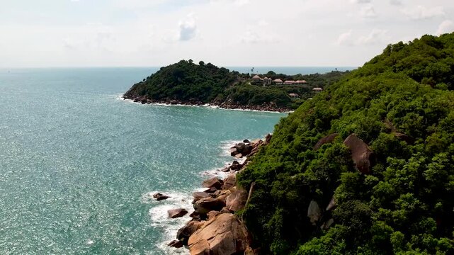A drone video of a coastline with turquoise water and rocky cliffs in Koh Samui, Thailand, stretching under a bright  sky 