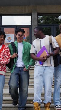 Vertical. Diverse group of smiling male and female university students walking down the stairs together after class, chatting and laughing while carrying books and backpacks on campus grounds