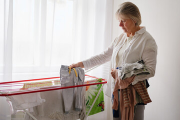 A senior woman carefully hangs freshly washed clothes on a drying rack in a sunny indoor setting,...