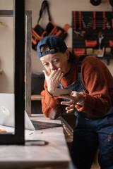 Worried young female mechanic in coveralls holding credit card and looking upset at laptop screen in her garage workshop.