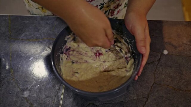 Close-up of hands diligently mixing a creamy batter with fresh blueberries in a grey bowl. This captures the essence of preparing homemade baked goods, promising a delicious and fruity treat.