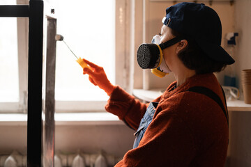 Side view woman worker in respirator applying brush paint metal table frame furniture restoration...