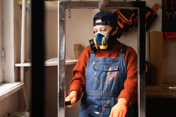 Female worker in coveralls and respirator painting metal construction with a brush inside home garage