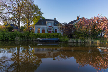 Traditional Dutch house by a canal in Bedum, Netherlands, with bright red windows and door. Boat, blossoming tree, and reflection in the water in warm evening light