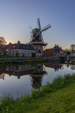 Traditional windmill De Vlijt in Bedum, Netherlands, silhouetted at sunset. The mill and surrounding brick houses reflect in the calm canal water