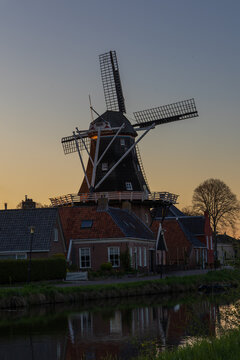Traditional windmill De Vlijt in Bedum, Netherlands, at dusk. The illuminated mill stands above brick houses and reflects in the calm canal water