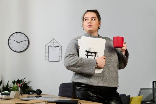Confident plus size woman holding papers and a cup in a stylish modern workspace - Powered by Adobe