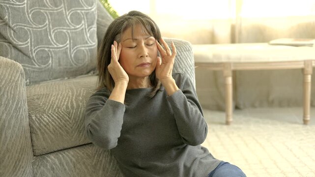 Mature woman suffering headache stress pain rubbing face sitting on living room floor bright window light background