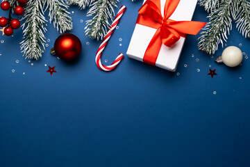 Christmas present box, white and red decorations and fir tree branches at blue background. Flat lay image with space for design.