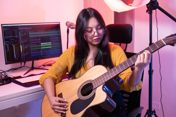 A female guitarist plays acoustic guitar indoors in a personal workspace, showing focus, creativity, and enjoyment of music during a quiet practice session.