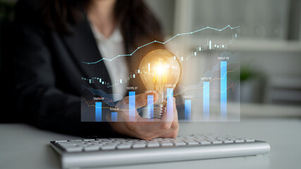 A business professional demonstrates innovation by holding a glowing light bulb over a keyboard. Behind them, graphic financial data shows trends and growth in a modern office setting. Scalp