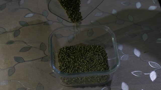 green moong beans being poured into a glass container on a patterned table