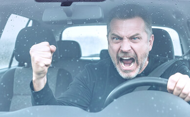 Homme d'&acirc;ge moyen furieux et agressif conduisant une voiture, criant avec le poing lev&eacute;, vu &agrave; travers un pare-brise mouill&eacute; par la pluie