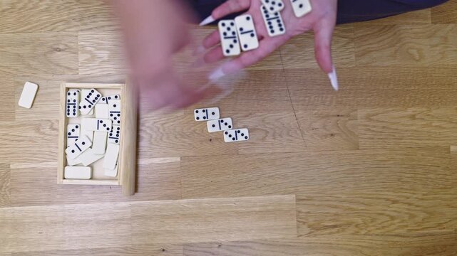 Close-up view of a person's hands carefully selecting and placing domino tiles on a light wooden floor, engaging in a relaxing and strategic game.
