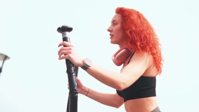 Fit redhead woman in sportswear using stepper machine for cardio workout, focused and determined with headphones on bright outdoor setting