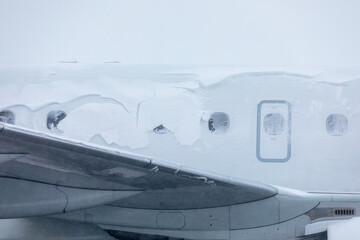 Snow covered airplane windows wait for deicing during cold winter day at airport. Scene shows frosty conditions on aircraft surface creating typical icy atmosphere of winter operations.