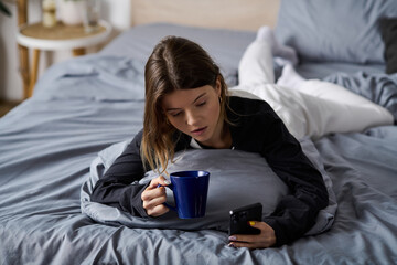 Young woman enjoys a cozy day in bed with coffee and her phone in a modern apartment