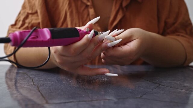 Close-up on a woman's hands as she uses a specialized electric nail file to meticulously shape and refine her long, white acrylic nails, performing a detailed manicure.