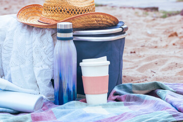 Beach picnic items with a straw hat, reusable water bottle, coffee cup, and cooler bag on a plaid blanket, enjoying outdoor leisure and eco friendly travel