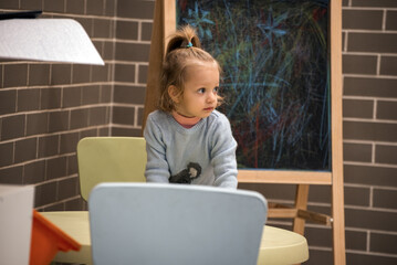 A little girl is sitting at a table in a room with a chalkboard on the wall