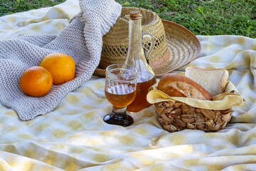 Picnic spread on yellow and white checkered blanket with oranges, bread basket, straw hat, bottle and glass, knitted throw for a relaxed outdoor summer al fresco moment