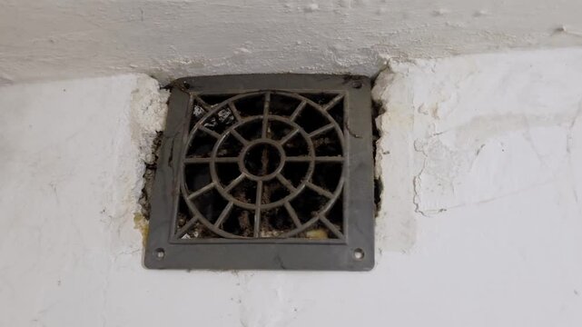 Low angle close-up shot of a dusty grey ventilation grille mounted on a rough white wall. The circular patterned vent is clogged with thick dust, cobwebs, and grime, indicating a lack of cleaning and