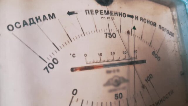 Macro shot of a vintage analog barometer dial face featuring Cyrillic text markings. The white gauge displays atmospheric pressure numbers and a thermometer scale.