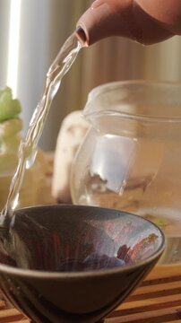 Close-up of boiling water being poured from clay teapot into glass pitcher and ceramic tea bowl set on wooden tea tray. Warming up tea brewing utensils. Traditional chinese gongfu tea ceremony at home
