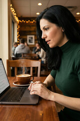 A woman is sitting at a table with a laptop and a cup of coffee. She is typing on the laptop and she is focused on her work. The scene suggests a quiet and productive atmosphere
